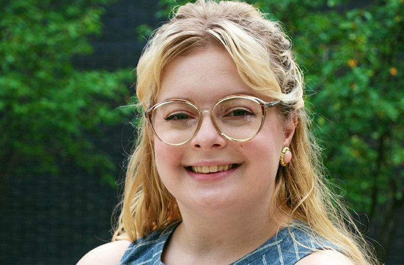 Smiling woman with glasses and blonde hair, wearing a sleeveless top, standing outdoors with greenery in the background.